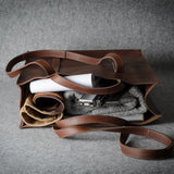A brown leather tote bag with a long strap, shown open and filled with rolled garments, a white tube, and a gray textured item, set against a gray fabric background.