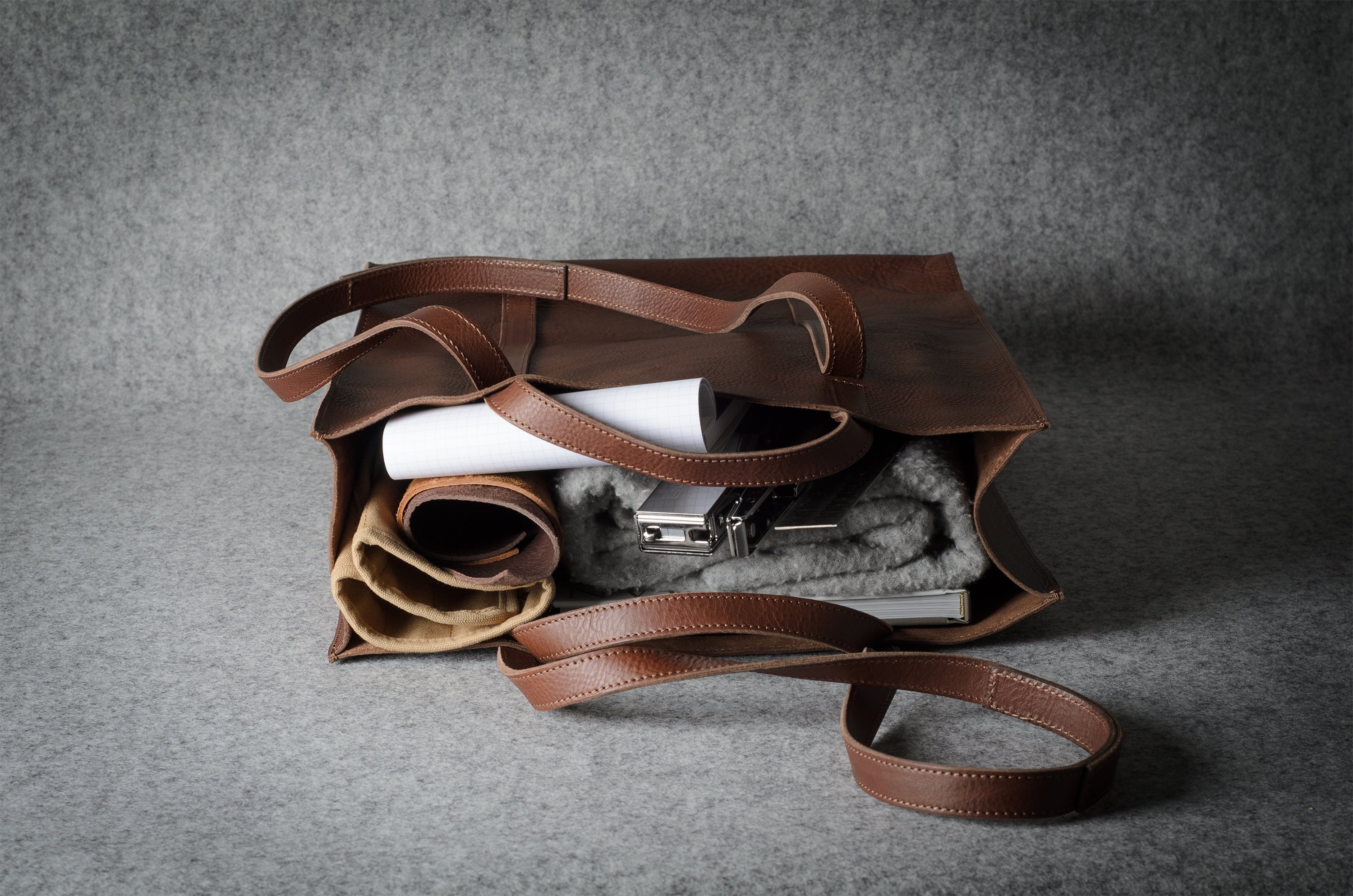 A brown leather tote bag with a long strap, shown open and filled with rolled garments, a white tube, and a gray textured item, set against a gray fabric background.