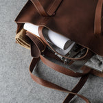 A brown pebbled leather tote bag shown from an overhead angle, partially open to reveal a white cylindrical item, a beige fabric piece, and a gray textured insert inside, set against a gray felt background.