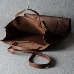 A brown leather tote bag with a matching shoulder strap, shown open against a textured gray background.