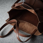A close-up of a brown leather tote bag shown open, highlighting its dual-texture exterior and interior lining on a gray textured surface.