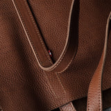 Close-up of a brown pebbled leather tote bag showing the texture, stitching, and a small red brand tab on the strap.
