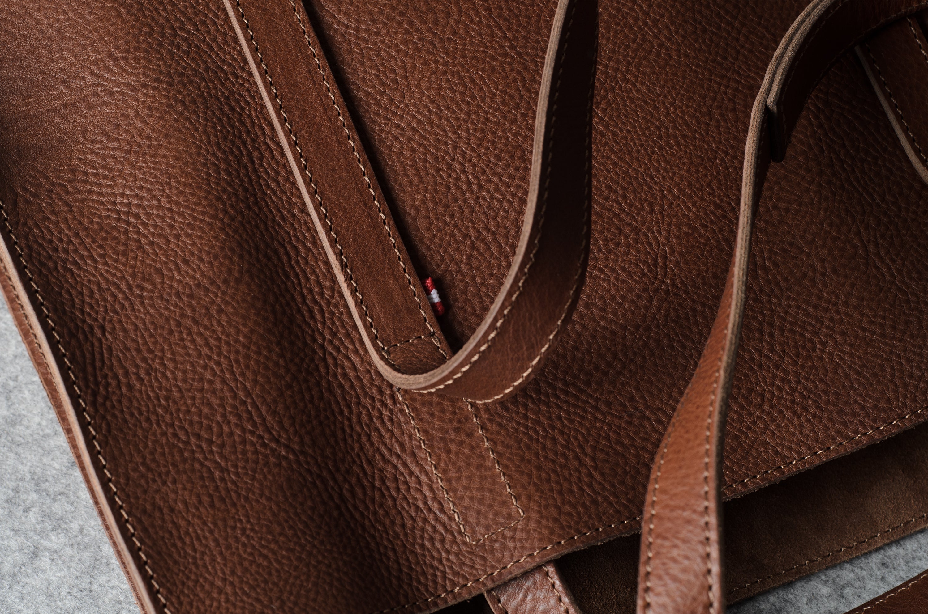 Close-up of a brown pebbled leather tote bag showing the texture, stitching, and a small red brand tab on the strap.