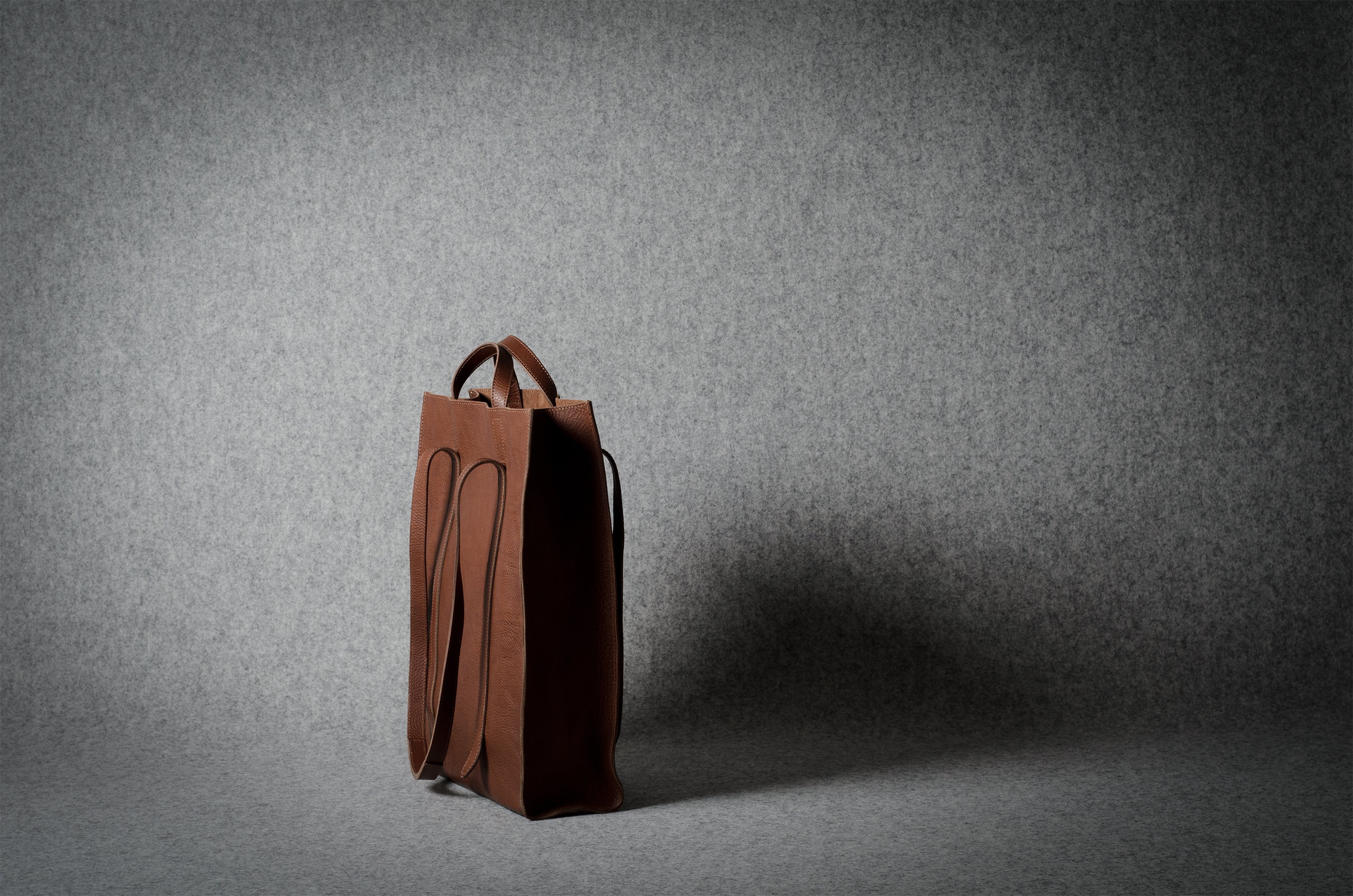 A brown leather tote bag with integrated handles, standing upright on a gray textured studio floor against a matching gray backdrop.