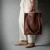 A person wearing a cream-colored outfit and tan shoes holds a large brown leather tote bag against a dark gray textured studio background.
