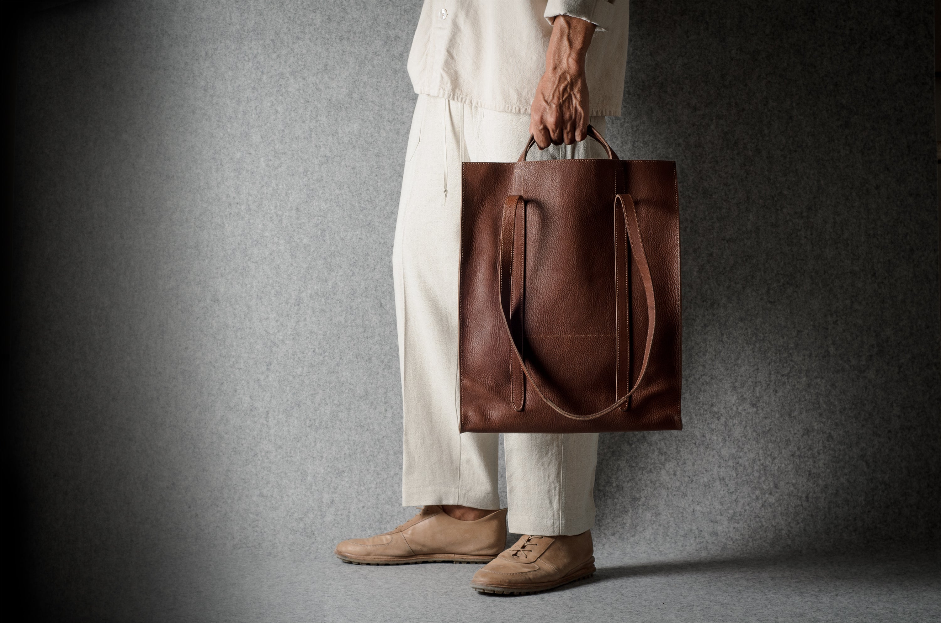 A person wearing a cream-colored outfit and tan shoes holds a large brown leather tote bag against a dark gray textured studio background.