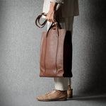 A person in a beige blazer and white trousers holds a large brown leather tote bag against a dark gray textured studio backdrop.