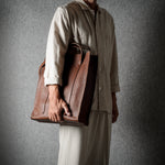 A person wearing a light beige traditional outfit holds a large brown leather tote bag against a dark gray textured background.