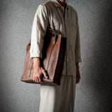 A person wearing a light beige traditional outfit holds a large brown leather tote bag against a dark gray textured background.
