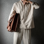 A person wearing a light beige button-up shirt and matching trousers, holding a brown leather tote bag against a dark textured gray background.