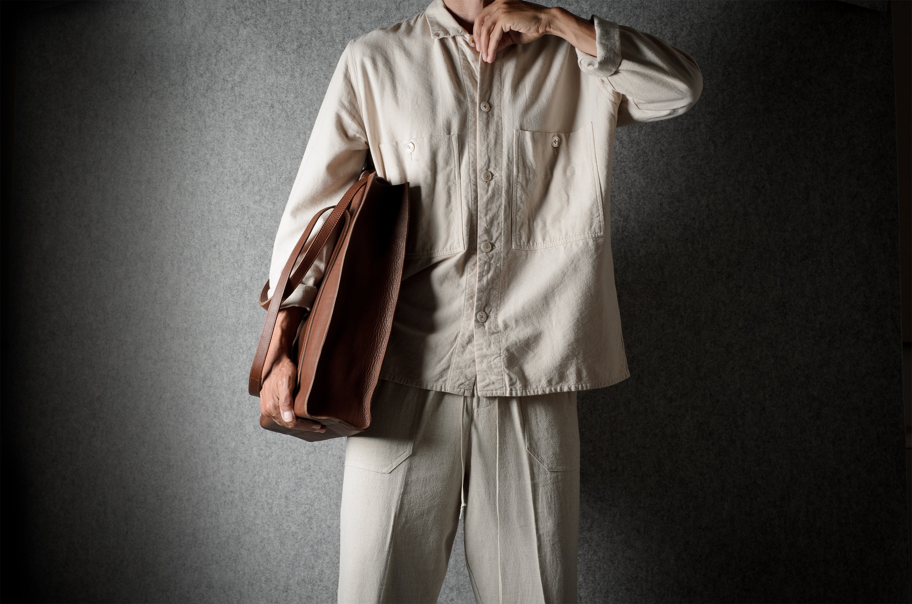 A person wearing a light beige button-up shirt and matching trousers, holding a brown leather tote bag against a dark textured gray background.
