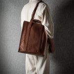 A person wearing a cream-colored outfit is shown from behind, carrying a large brown leather tote bag over their shoulder against a dark textured gray wall.
