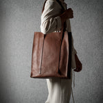 A person in a cream-colored outfit carries a large brown leather tote bag against a textured gray background.