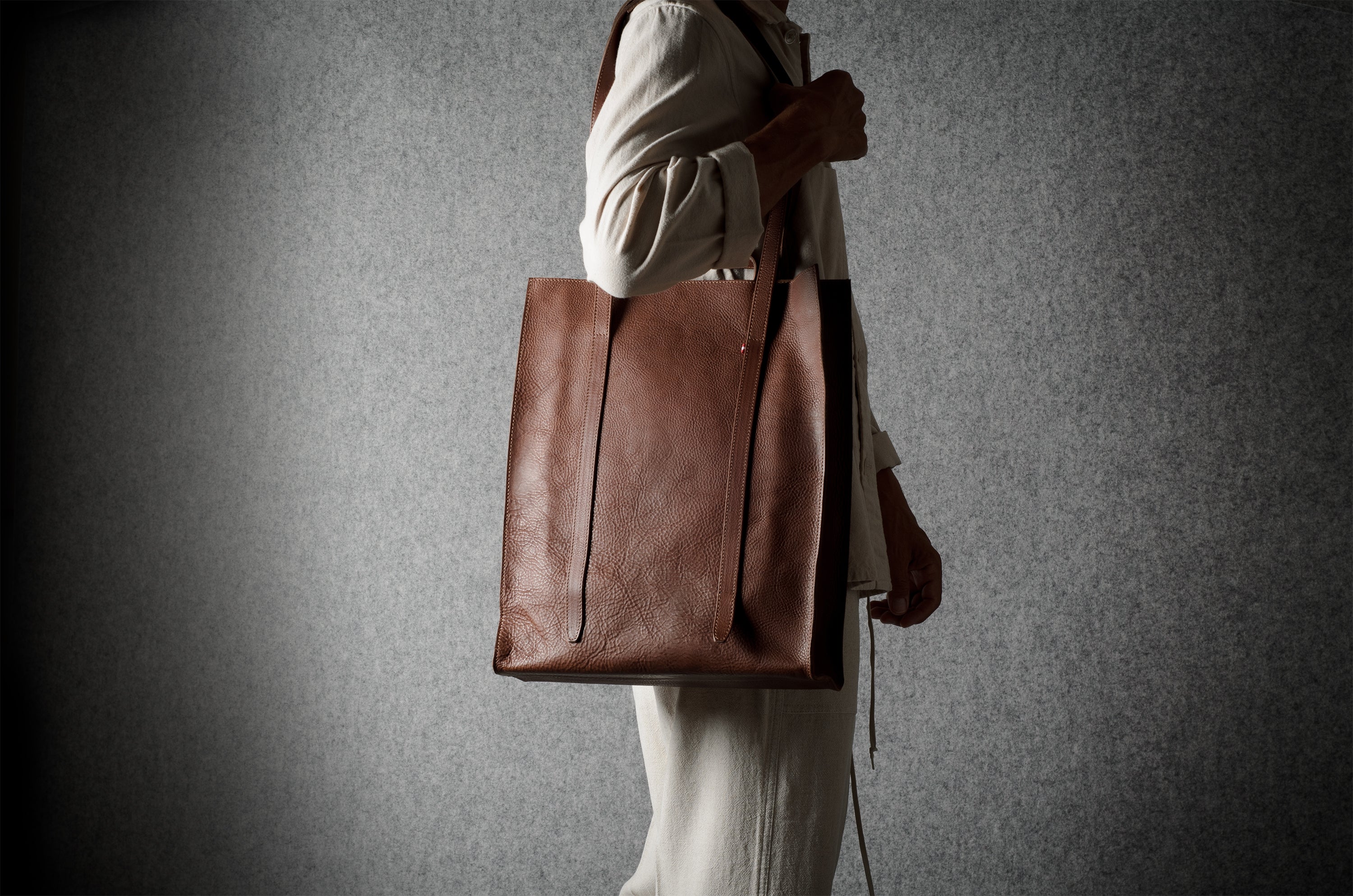 A person in a cream-colored outfit carries a large brown leather tote bag against a textured gray background.