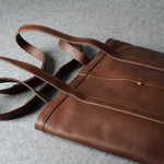 A brown leather tote bag with matching shoulder straps, laid flat on a textured gray surface.