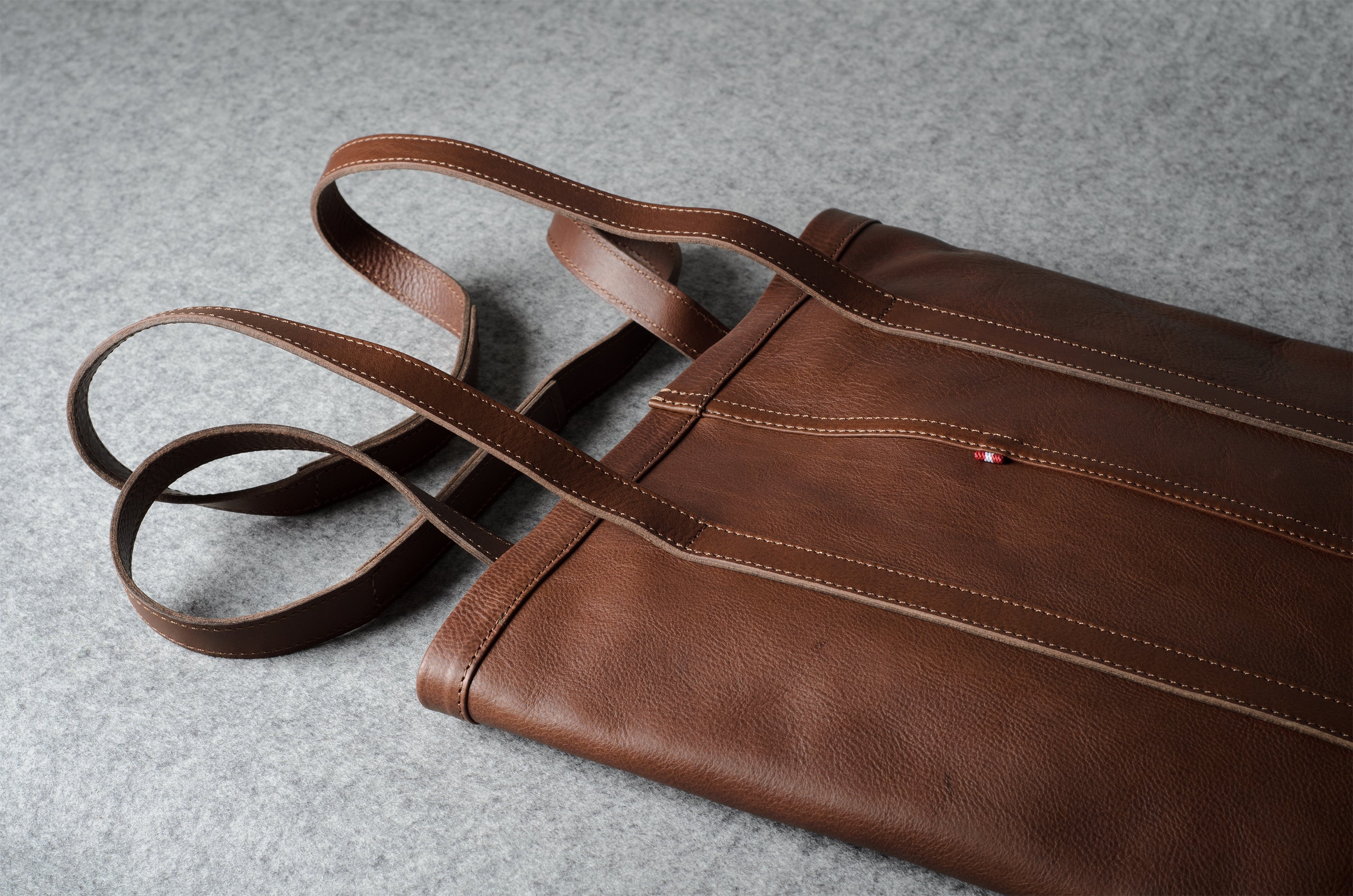 A brown leather tote bag with matching shoulder straps, laid flat on a textured gray surface.