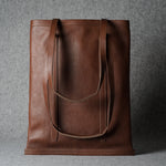 A brown leather tote bag with long shoulder straps, standing upright against a textured gray background.