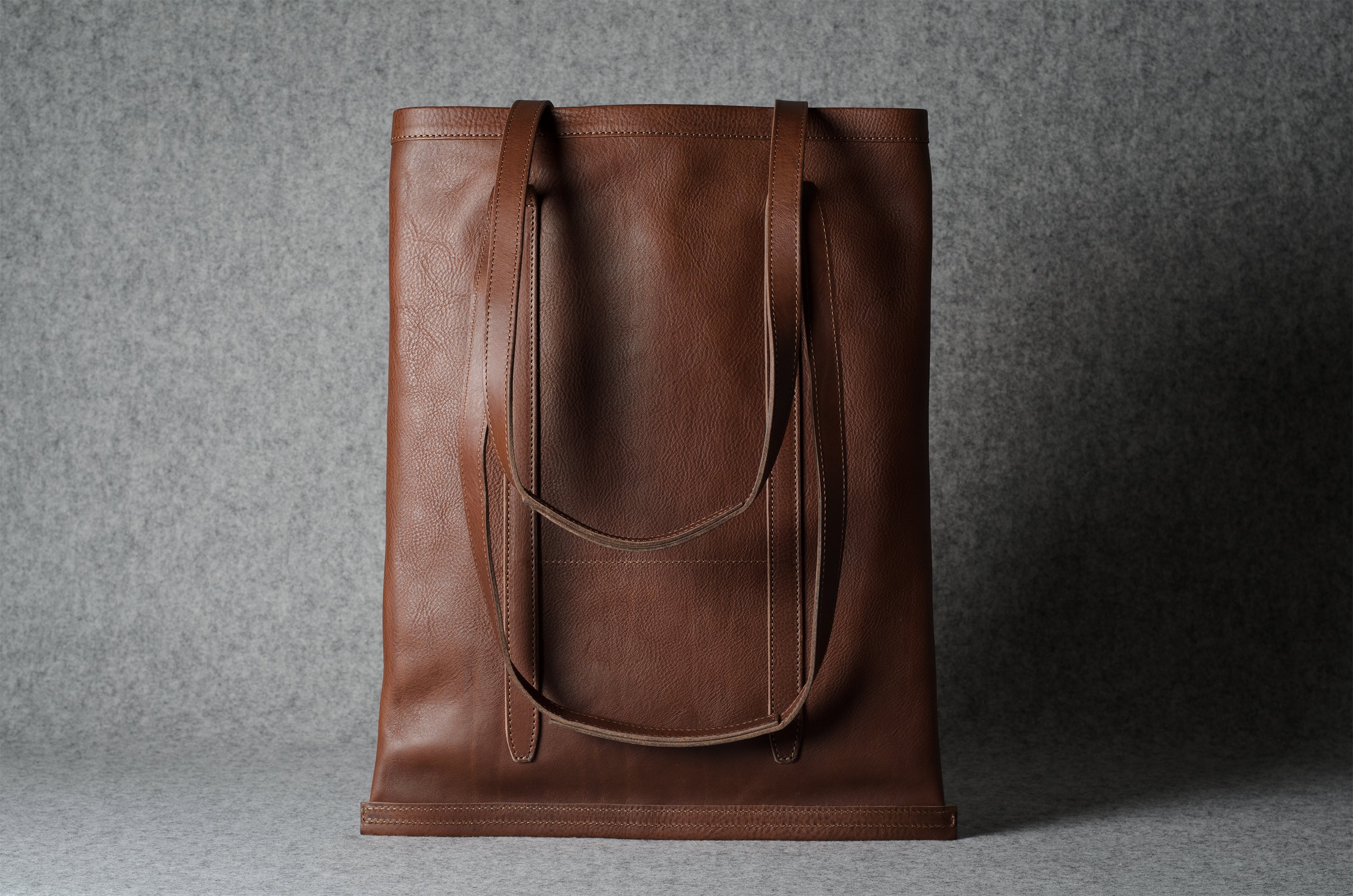 A brown leather tote bag with long shoulder straps, standing upright against a textured gray background.