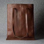 A brown leather tote bag with two long shoulder straps, standing upright against a textured gray background.