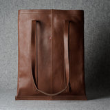 A brown leather tote bag with two long shoulder straps, standing upright against a textured gray background.