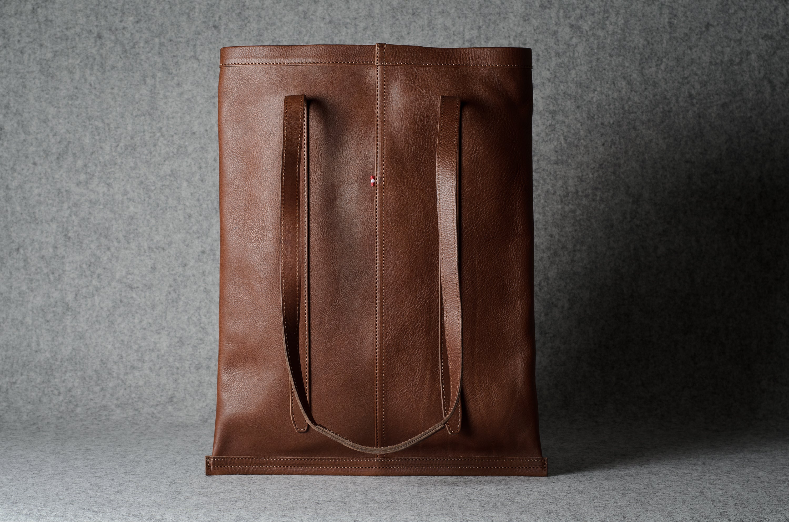 A brown leather tote bag with two long shoulder straps, standing upright against a textured gray background.