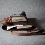 A brown leather tote bag with a long shoulder strap, shown open and styled with books, a notebook, and a small bottle on a textured gray background.