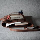 A brown leather tote bag with a long shoulder strap, shown open and styled with books, a notebook, and a small bottle on a textured gray background.