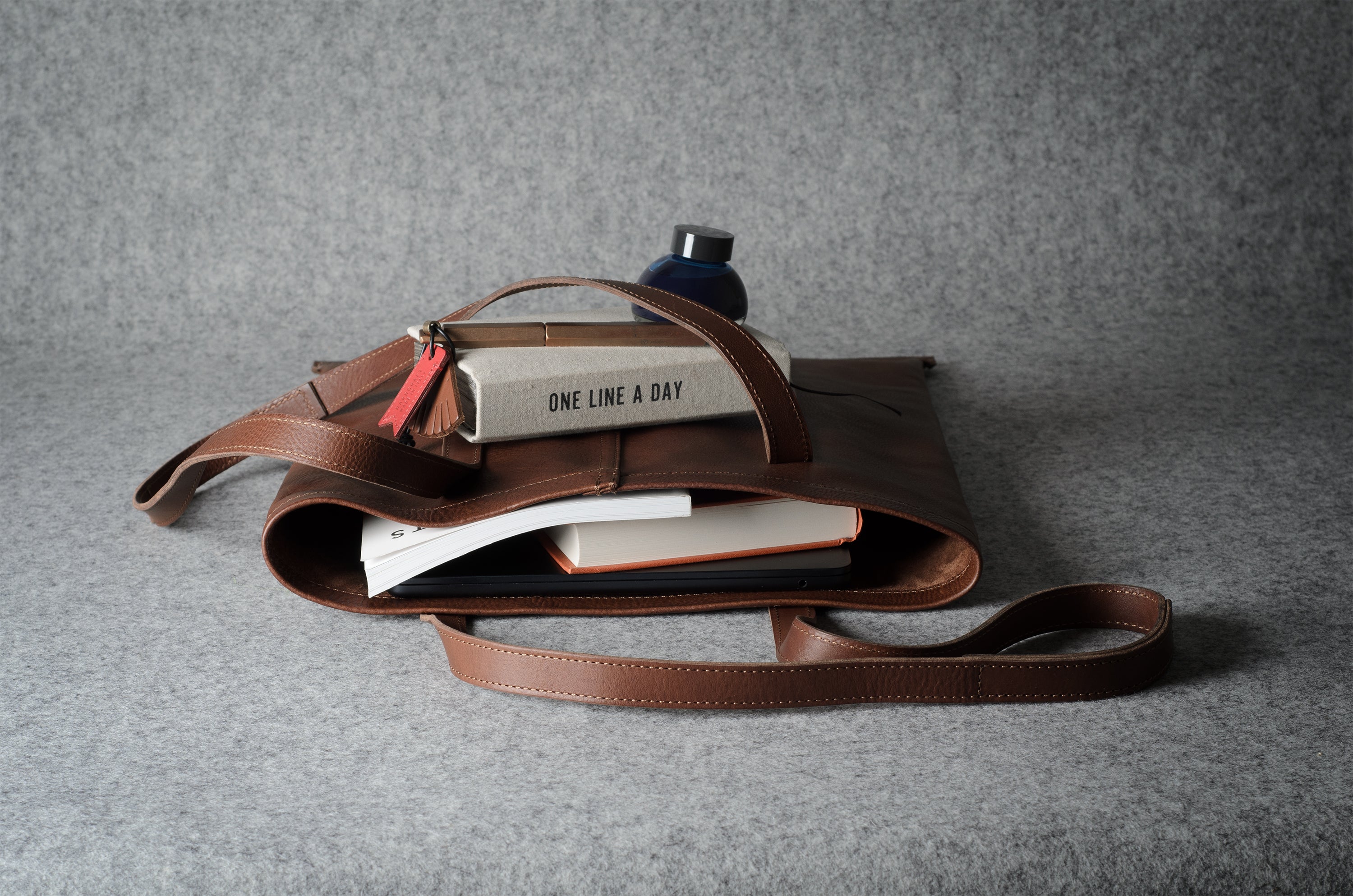 A brown leather tote bag with a long shoulder strap, shown open and styled with books, a notebook, and a small bottle on a textured gray background.