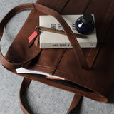 A brown leather tote bag shown from above on a gray textured surface, with a book, pen, and small bottle placed on top.