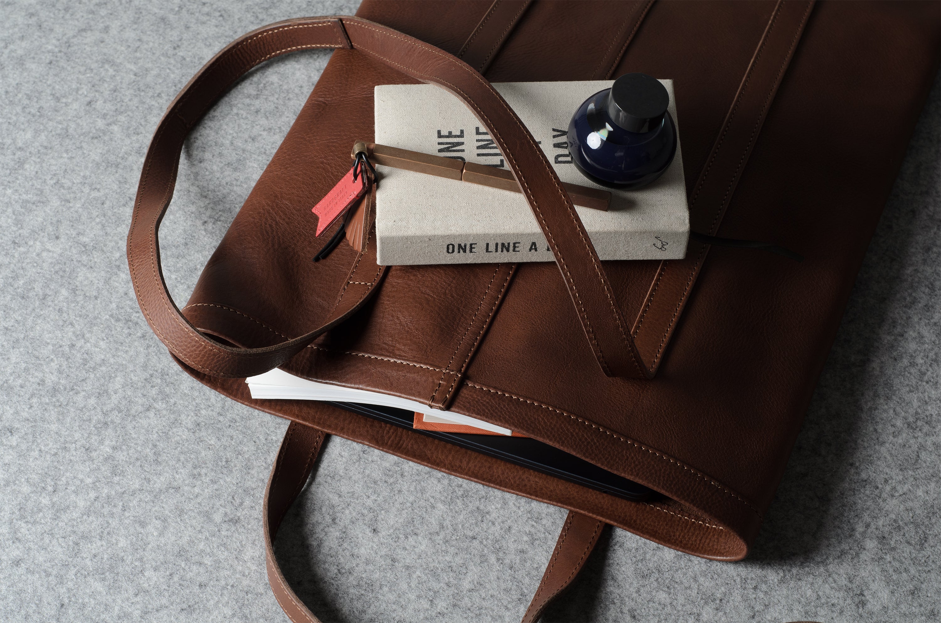 A brown leather tote bag shown from above on a gray textured surface, with a book, pen, and small bottle placed on top.