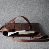 A brown leather tote bag with a long shoulder strap, shown open with books inside, set against a textured gray background.