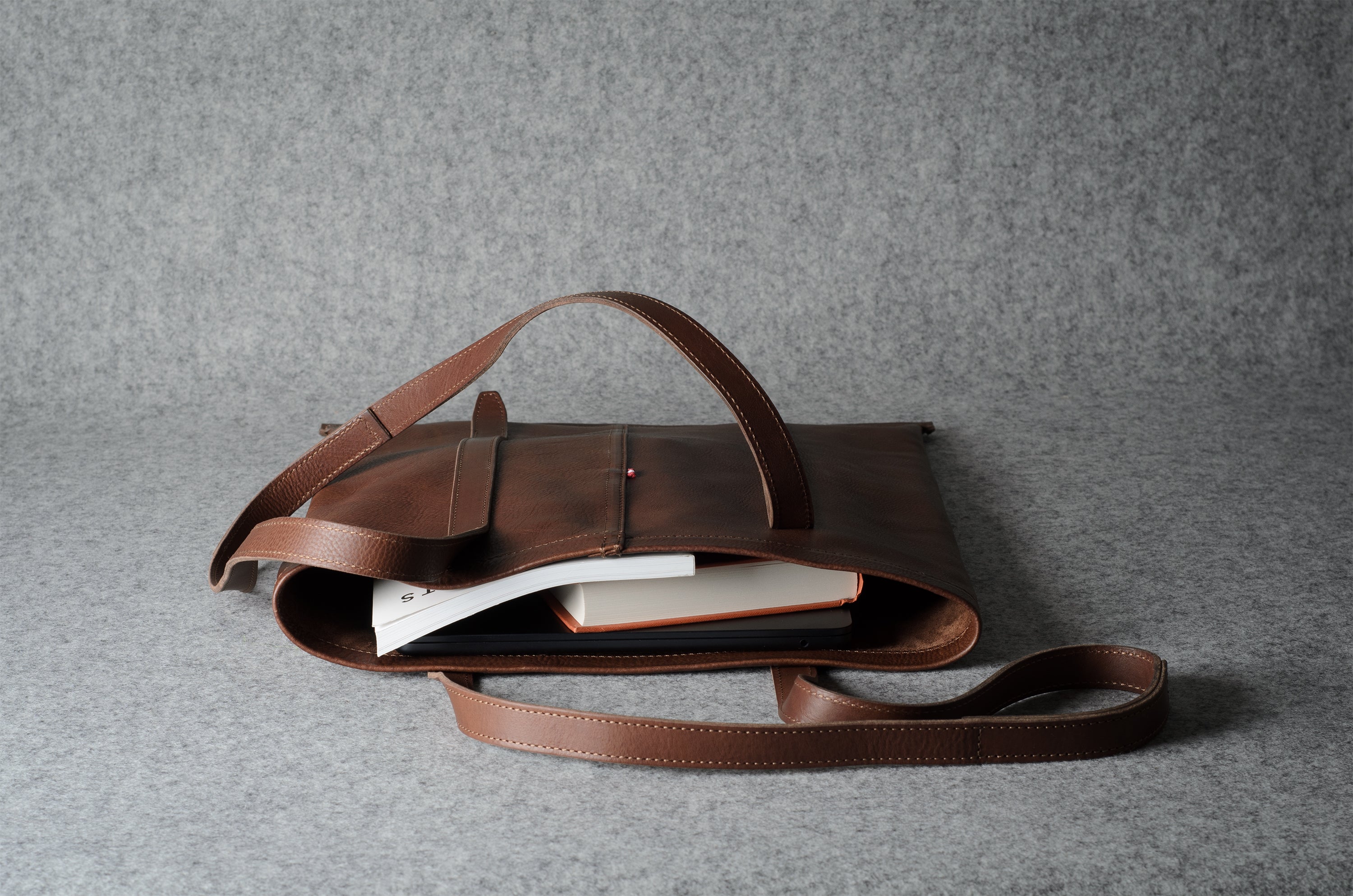 A brown leather tote bag with a long shoulder strap, shown open with books inside, set against a textured gray background.