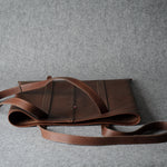 A brown leather shoulder bag with a long strap, displayed on a textured gray background.