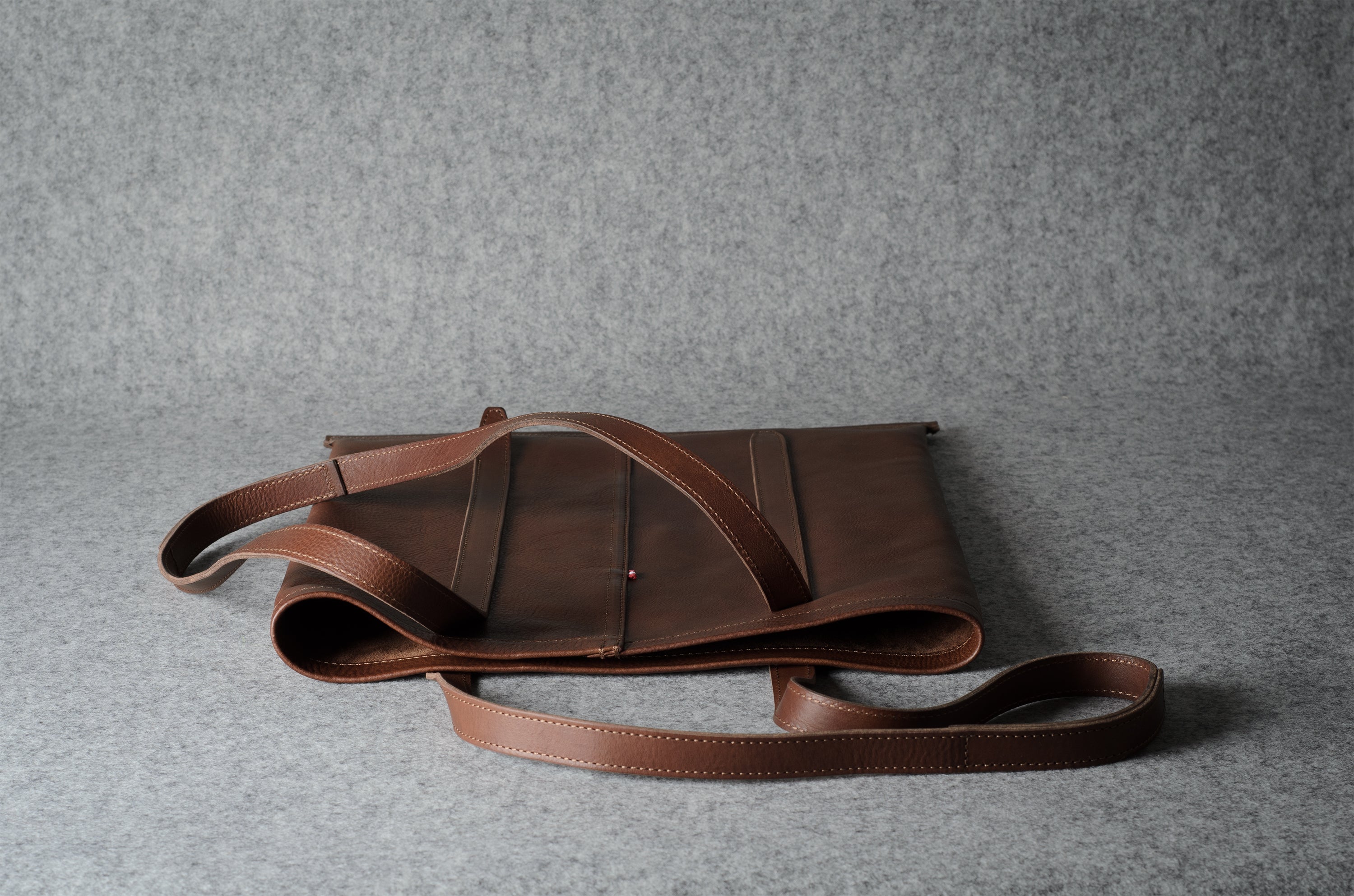 A brown leather shoulder bag with a long strap, displayed on a textured gray background.