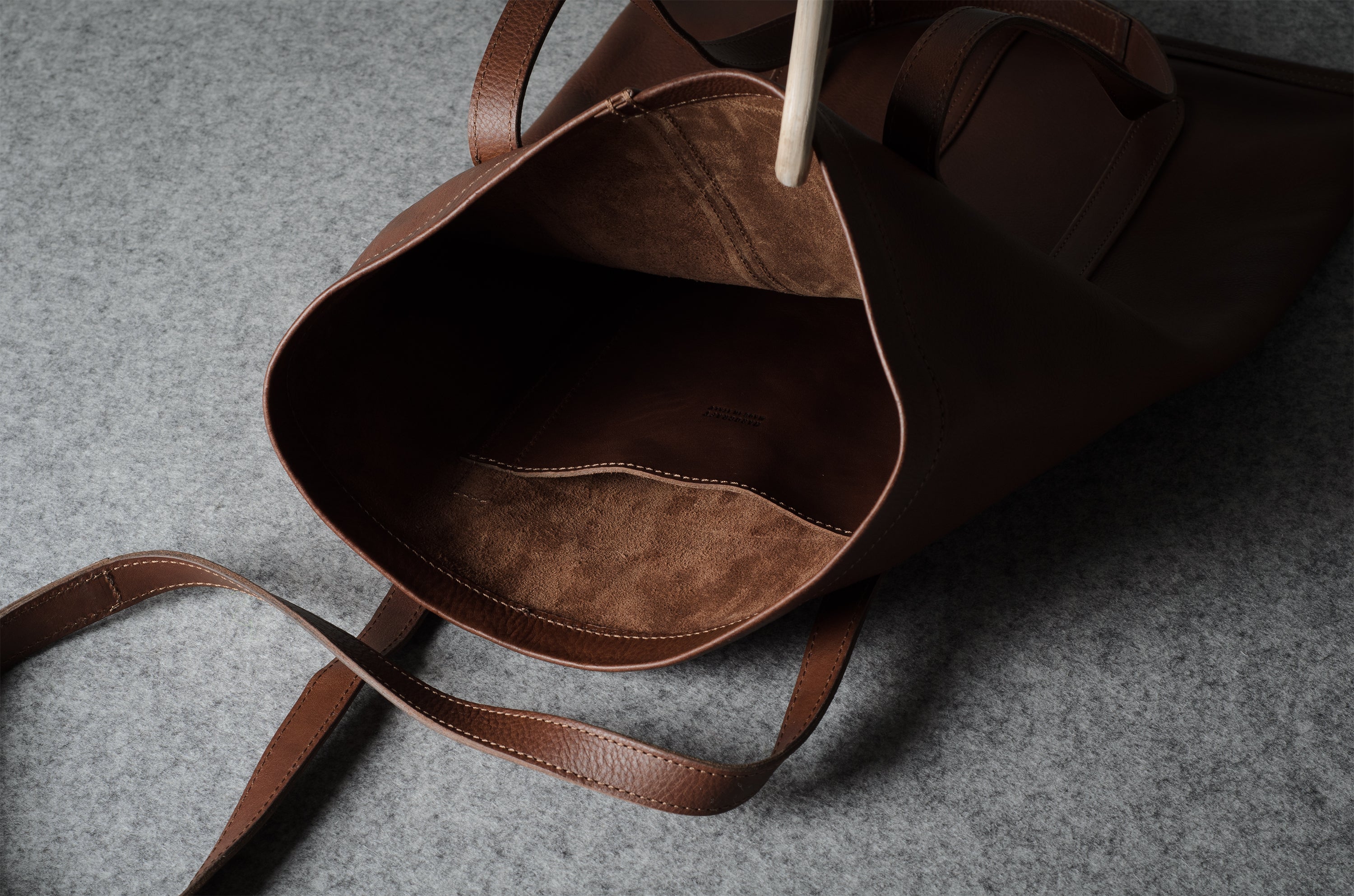 A close-up of a dark brown leather tote bag shown from the inside, highlighting the rich brown interior lining and matching shoulder straps on a textured gray surface.