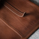 Close-up of a brown leather bag showing the shoulder strap, stitching, and a strap against a gray textured background.