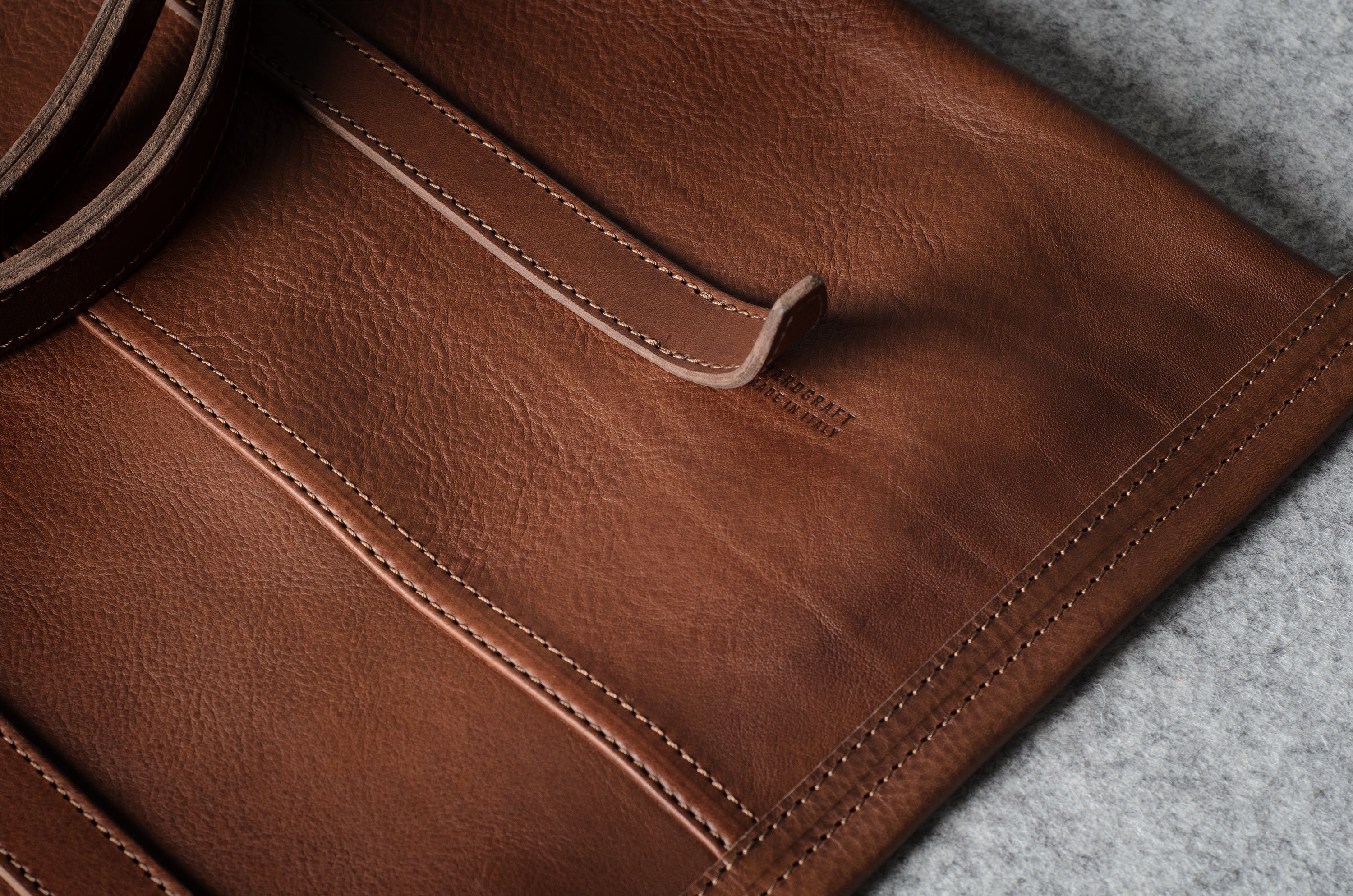 Close-up of a brown leather bag showing the shoulder strap, stitching, and a strap against a gray textured background.
