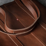 Close-up of a brown leather tote bag with matching shoulder straps, showing fine stitching and a small red brand tab.