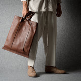 A person wearing light beige clothing and brown shoes holds a large brown leather tote bag against a dark gray textured studio background.