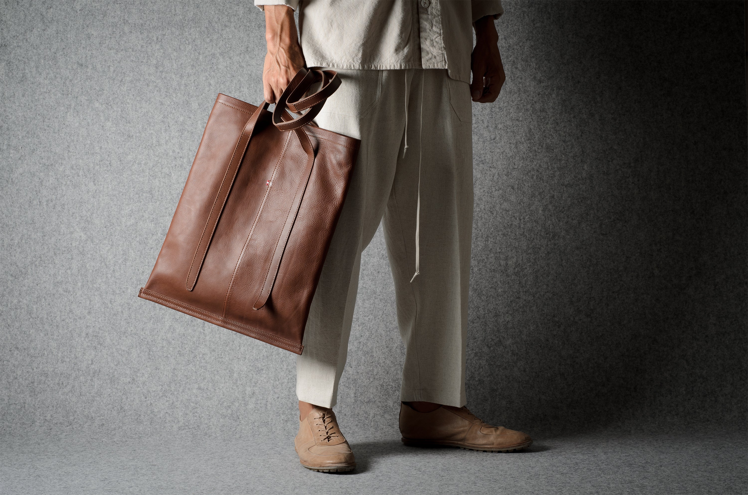 A person wearing light beige clothing and brown shoes holds a large brown leather tote bag against a dark gray textured studio background.