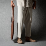 A person wearing loose off-white drawstring trousers and tan shoes, holding a brown leather bag against a dark gray studio backdrop.