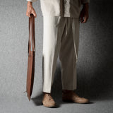 A person wearing loose off-white drawstring trousers and tan shoes, holding a brown leather bag against a dark gray studio backdrop.