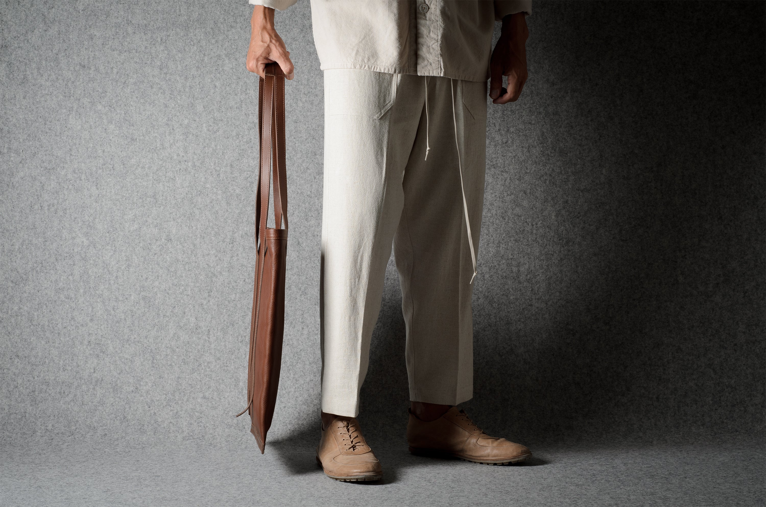 A person wearing loose off-white drawstring trousers and tan shoes, holding a brown leather bag against a dark gray studio backdrop.