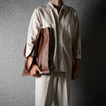 A person wearing a light beige button-up shirt and matching trousers holds a brown leather shoulder bag against a dark gray textured studio background.