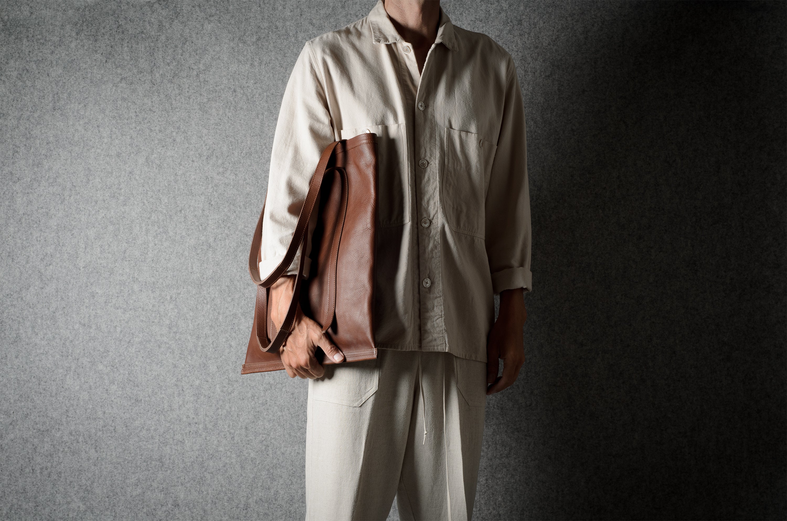 A person wearing a light beige button-up shirt and matching trousers holds a brown leather shoulder bag against a dark gray textured studio background.