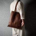 A person wearing a cream-colored suit is shown from the back, carrying a large brown leather tote bag over their shoulder against a textured gray wall.