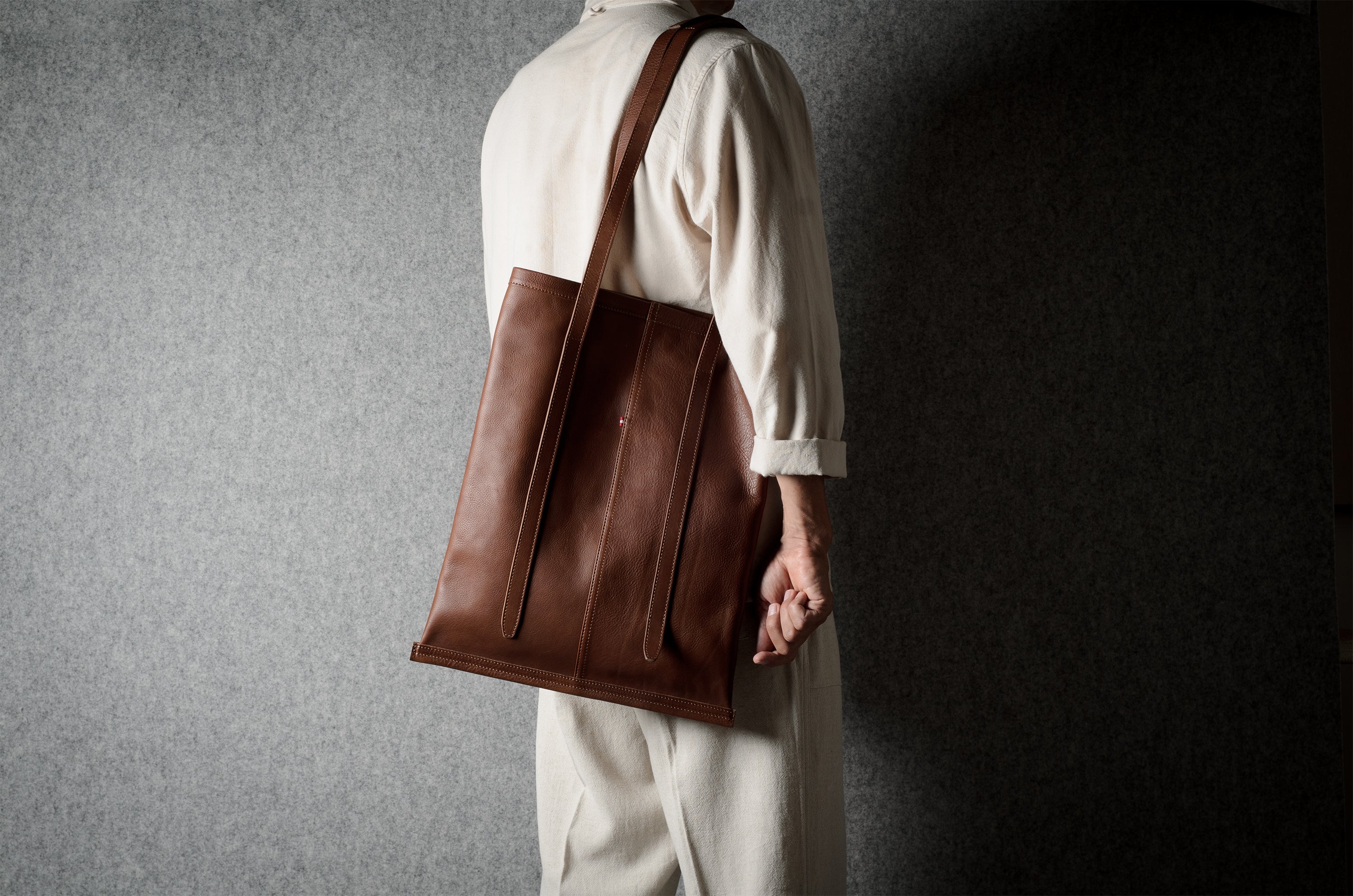 A person wearing a cream-colored suit is shown from the back, carrying a large brown leather tote bag over their shoulder against a textured gray wall.
