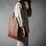 A person in a light beige shirt and trousers carries a large brown leather tote bag against a textured gray background.