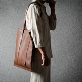 A person in a light beige shirt and trousers carries a large brown leather tote bag against a textured gray background.