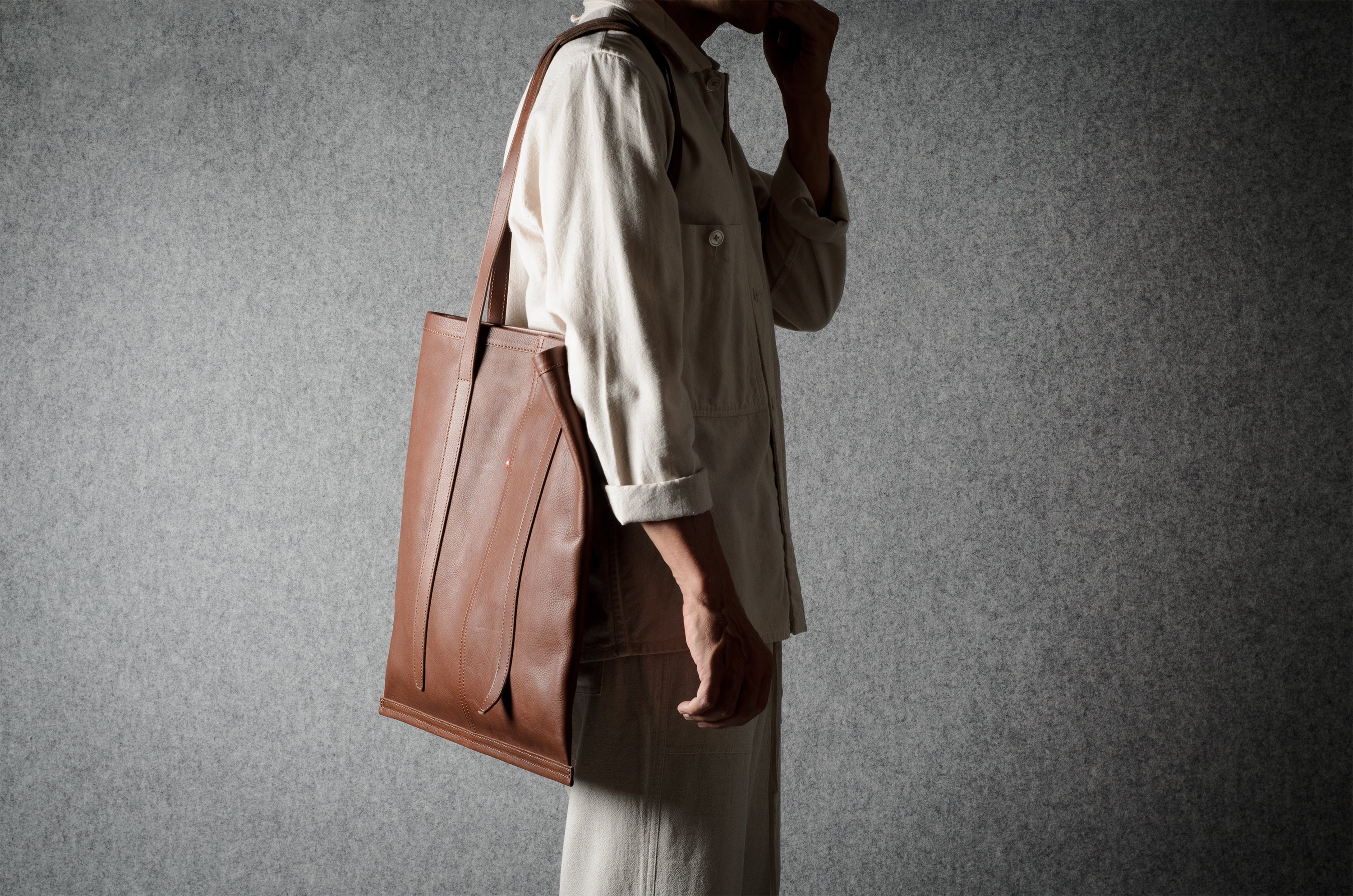 A person in a light beige shirt and trousers carries a large brown leather tote bag against a textured gray background.